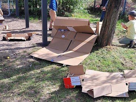 Kinder sammeln Bastelmaterialien auf dem Spielplatz. Daneben liegen angefangene Bastelprojekte aus großen Pappkartons