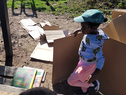 Ein Kind trägt eine große Pappe über den Spielplatz. Auf einer Bank lieben Bücher.