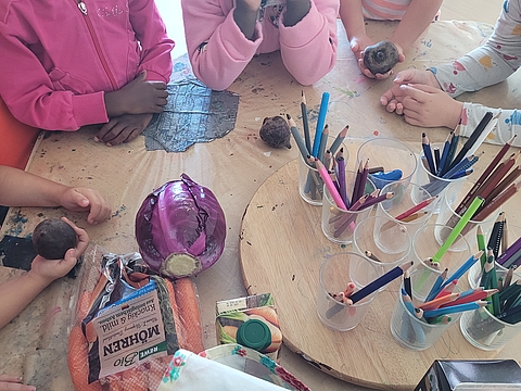 Kinder stehen um einen Tisch mit Buntstiften, einem Kohlkopf, Möhren und Rotebeete.