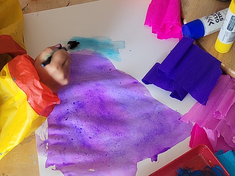 Eine Kinderhand bemalt ein Papier, daneben liegt buntes Krepppapier