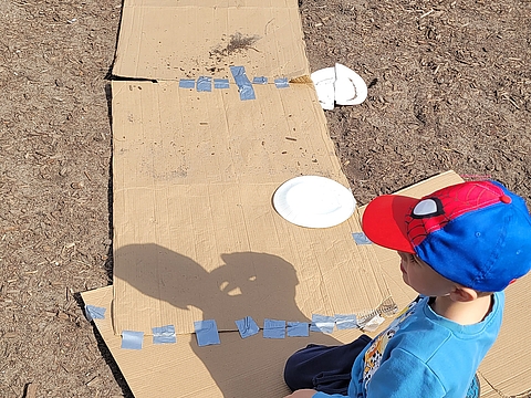 Mehrere Pappen liegen zusammengeklebt draußen auf dem Boden, darauf sitzt ein Kind.