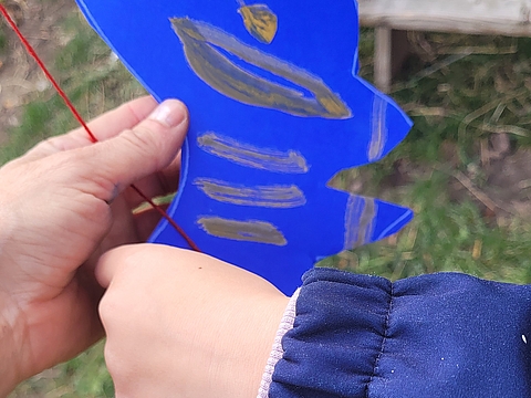 Eine erwachsene Hand hält eine bemalte Pappfigur während eine Kinderhand daran einen Faden befestigt