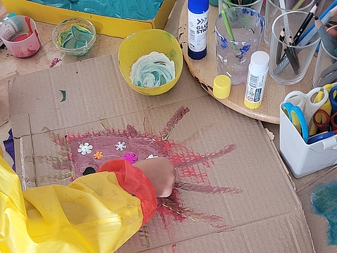 Eine Kinderhand klebt Dekoschneeflocken auf eine bemalte Pappe umgeben von Bastelutensilien