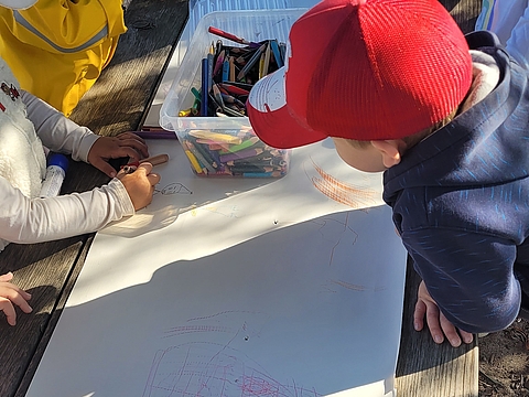 Eine Gruppe von Kindern malt mit Buntstiften auf einem mit Papier beklebten Tisch
